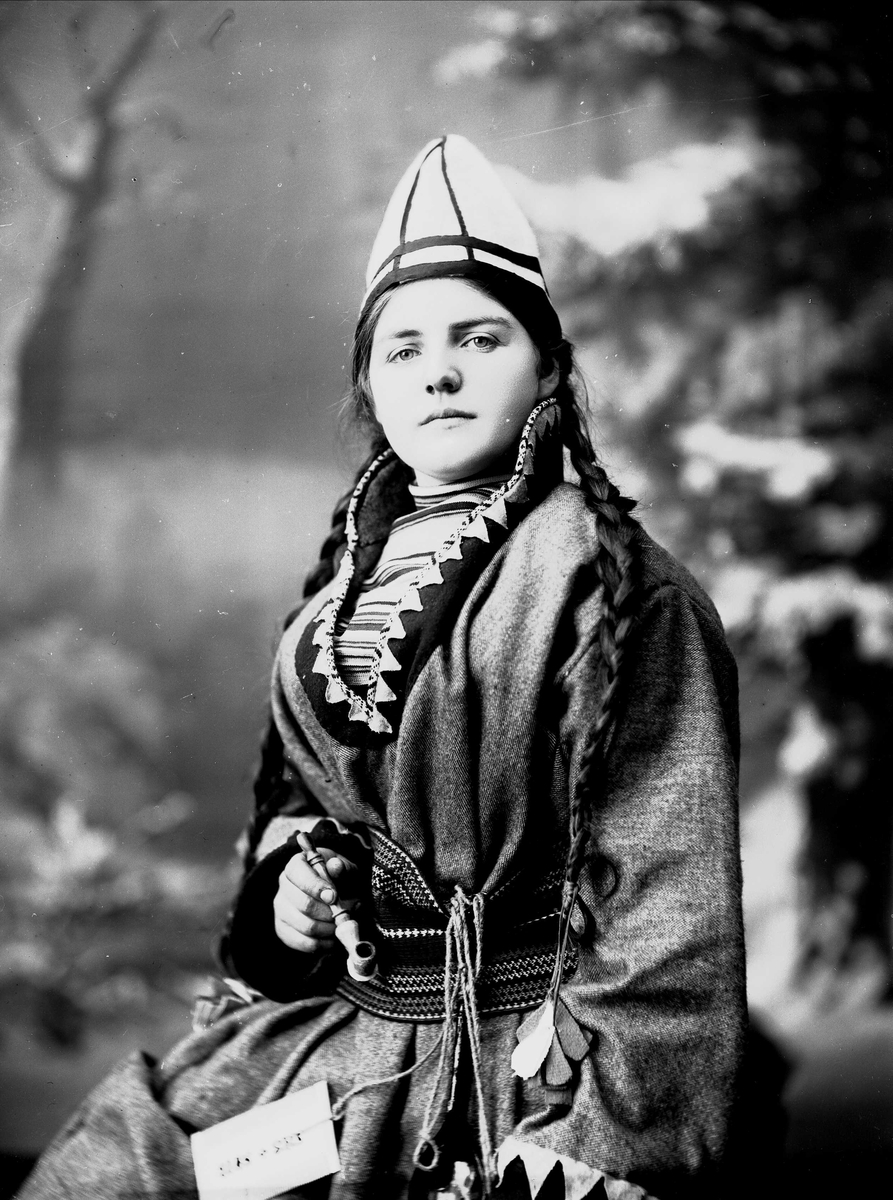 Solveig Lund, Kvinne i samisk drakt - kofte, lue og nålehus med beltering. I den ene hånden holder hun en pipe. Bakgrunnen er malt med trær.