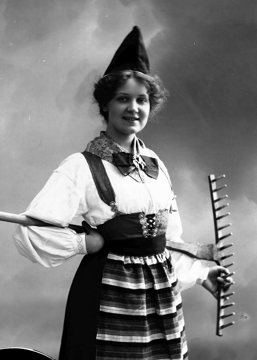 Solveig Lund, Kvinne i drakt og lue, som holder rive bak på ryggen.