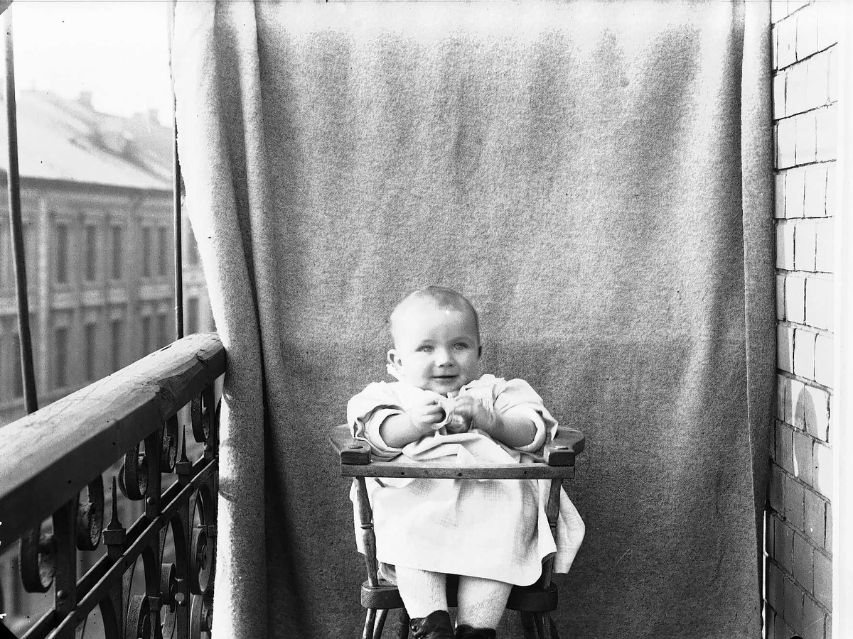 Axel Quinsgaard Wiborg, Lillebror Axel Q. Wiborg i barnestol på balkong, ant. Meltzers gate 9, Oslo, 1899. Fotografert mot opphengt bakteppe.