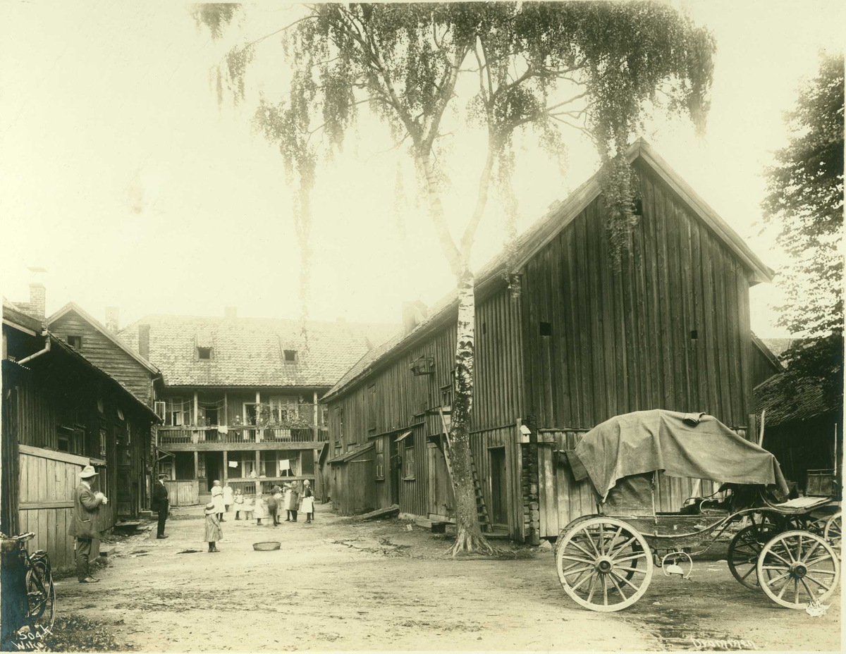 Anders Beer Wilse, Bygård, Drammen, Buskerud. Trebygninger rundt gårdsrom med barn og voksne. Uthus og bolig med svalgang. Hestekjøretøy plassert i forgrunnen.