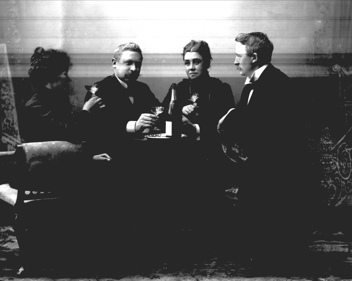 Gustav Borgen, Gruppeportrett, to herrer og to damer med champagne rundt stuebordet.