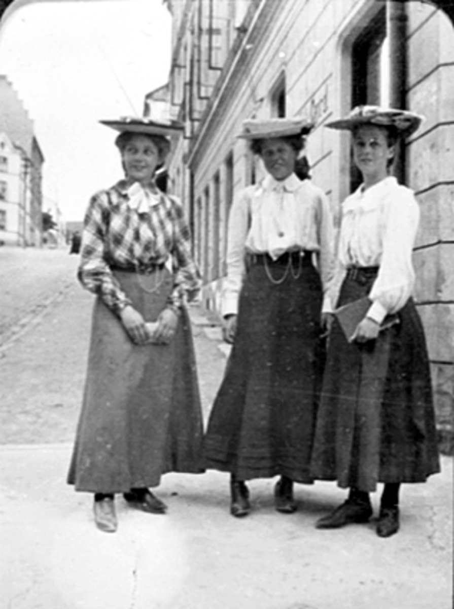 Anno Domkirkeodden, GRUPPE 3 UNGE DAMER MED HATT BLUSE OG FOTSIDE SKJØRT PÅ SPASERTUR, DET STÅR CENTRAL PÅ VEGGEN