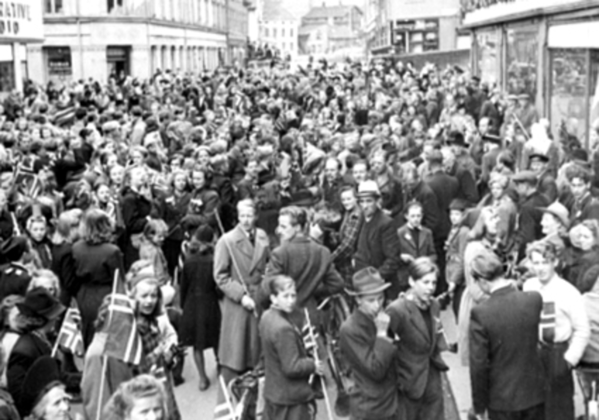 Anno Domkirkeodden, FREDSDAGENE I MAI 1945, TORGGATA, HAMAR. DA FREDEN VAR ET FAKTUM BLE DET MONTERT ET PAR KRAFTIGE HØYTALER UTENFOR RADIOFORRETNINGEN TIL O. OMMUNDSEN I FLAATERUDGÅRDEN, TORGGATA 43. FOLK STIMLET SAMMEN FOR Å HØRE NYHETER.