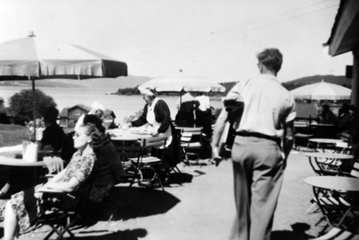 Anno Domkirkeodden, HEDMARKSMUSEET, DOMKIRKEODDEN, RESTUARANTEN, UTESERVERING, GJESTER, BETJENING, SOLPARASOLL. MED RYGGEN TIL, TEGNEREN PER SOLHEIM, SERVERINGSDAME ELINE MOEN, FØDT JOHANNESSEN, BRUMUNDDAL, F-1900-D. 1981. BILDET ER FRA FØRSTE HALVDEL AV 1950-TALLET.