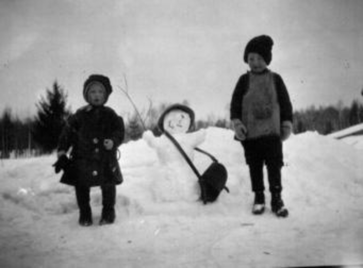 Anno Domkirkeodden, To barn leker i snøen. Odlaug Dobloug og Arne Dobloug. Snømann med skulderveske og hatt.