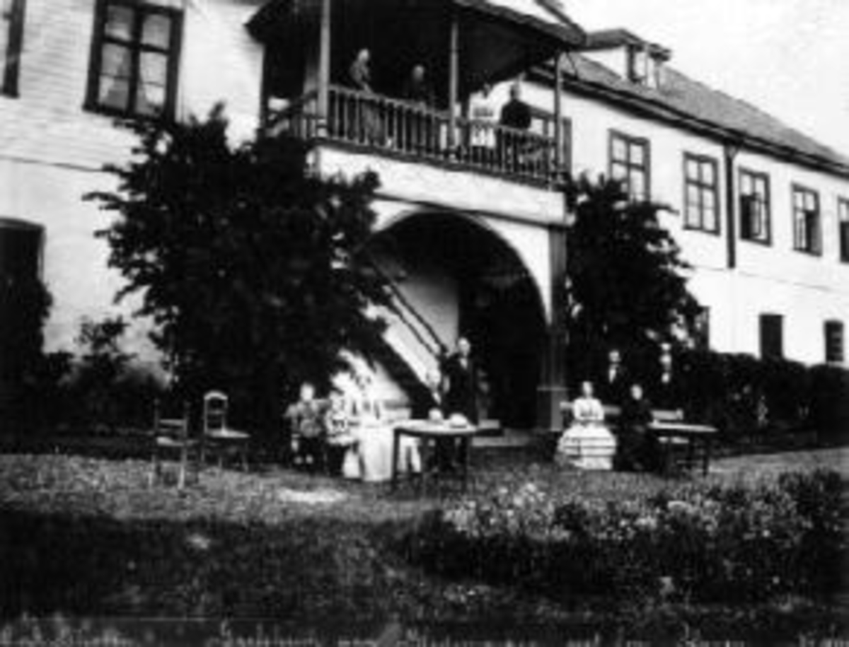 Anno Domkirkeodden, Eksteriør, hovedbygningen på Grefsheim, Nes, Hedmark. Fra hagen med bord, benker og stoler. Ved bordet er sorenskriver Egeberg med frue. Damen med stripet skjørt er Sophie Mellby.