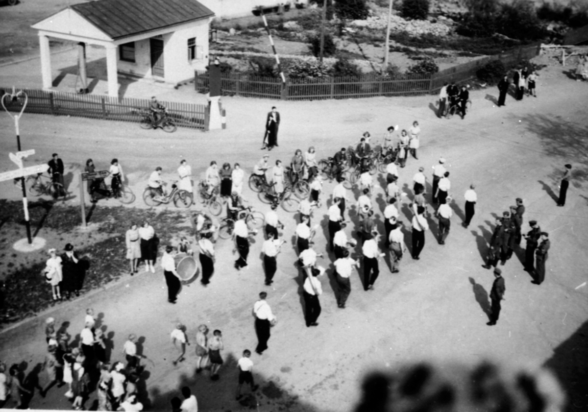 Anno Domkirkeodden, Brumunddal musikkorps marsjerer i Furnesvegen, Brumunddal. Bro mekaniske verksted bak til venstre. Tilskuere, også noen soldater (tyske?) til høyre.