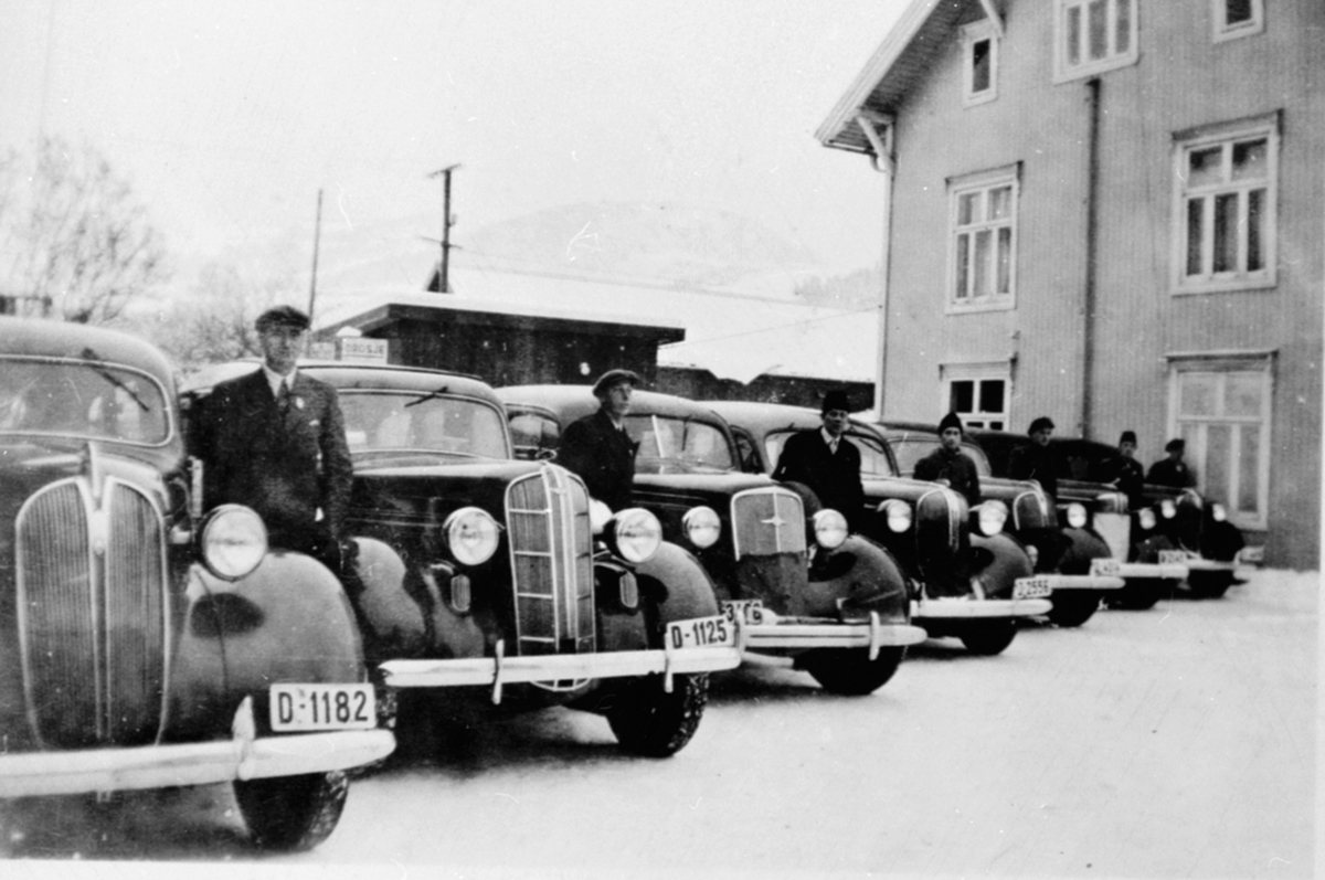 Anno Domkirkeodden, 7 drosjer med sjåfører oppstillt. Fra venstre er Martin Sørum med D-1182 Plymouth 1937, D-1125 Dodge 1936, Johan Lindbak med Chevrolet 1936, Olaf Stigen D-2565 Plymouth 1938, Guttorm Lehne D-4114 Plymouth 1937, Johannes Røseth D-2145 Dodge 1938, Johannes Lillesveen D-4414 Plymouth 1937, Mikael Bakk…