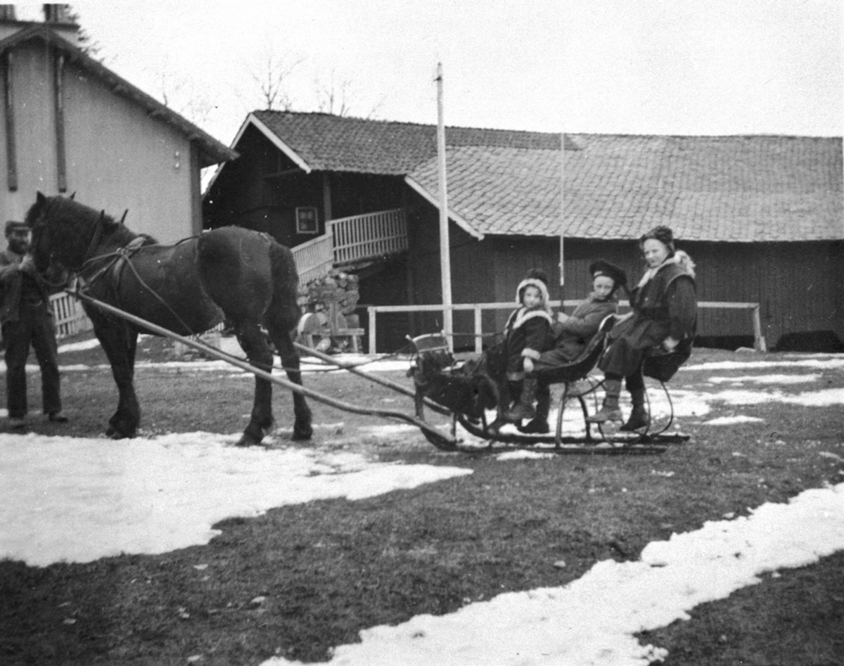 Anno Domkirkeodden, Hest med slede og tre barn på tunet til Nederkvern, Brumunddal. Fra venstre er Lily, Thore og Ingeborg Bjerke. Stallkar Smeby holder hesten. Stabburet og låven bak.
