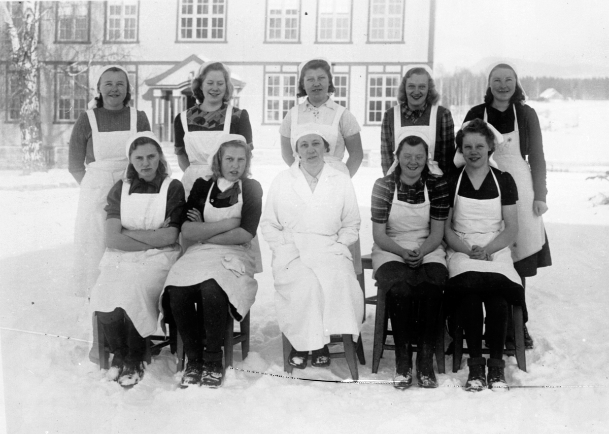 Anno Domkirkeodden, Husmorskoleelever med lærer ved Mørkved skole, Brumunddal. Utendørs med forklær og skaut. Vinter.