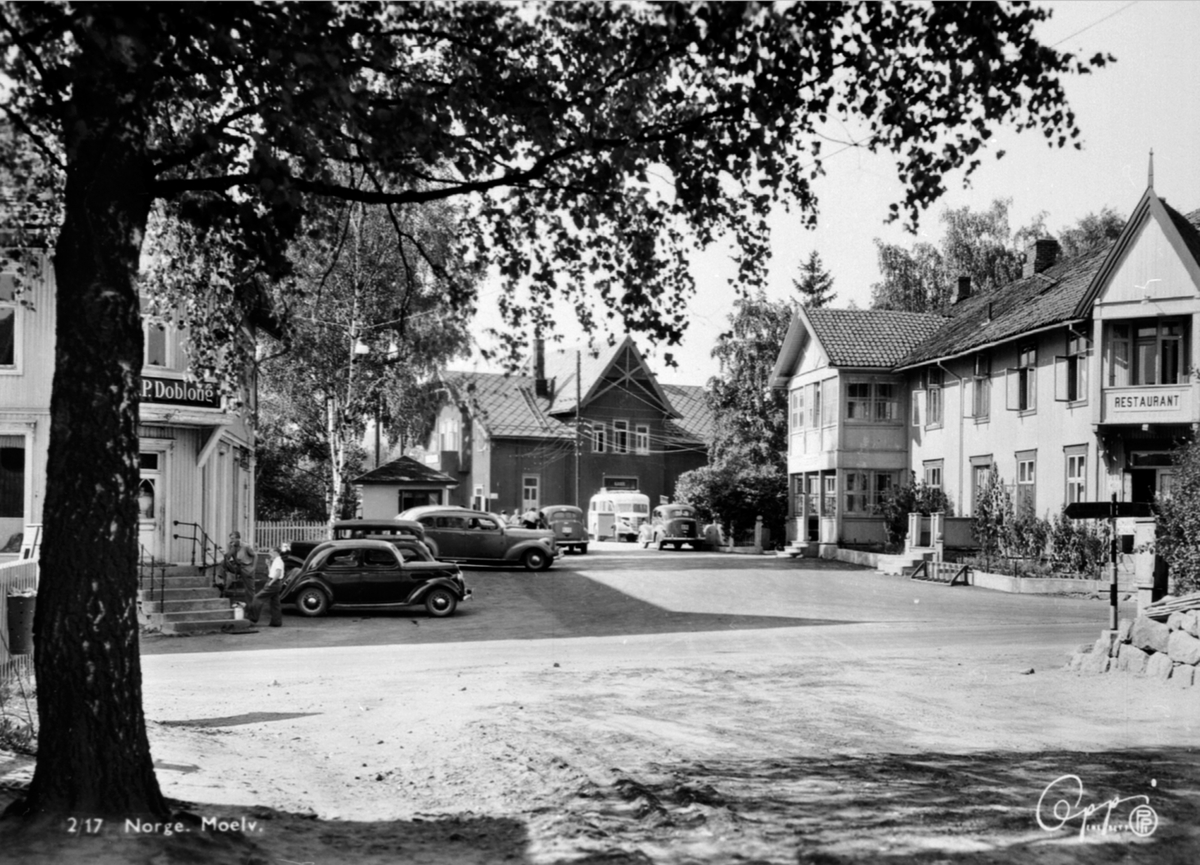 Anno Domkirkeodden, Krysset Åsmarkvegen, Storgata, Moelv. M.P. Dobloug, drosjebua, Moelv stasjon, Moelven Hotell, buss, biler, folk. Nærmest en fransk- evt. engelskbygd Ford V8 60 hk 1937-39.