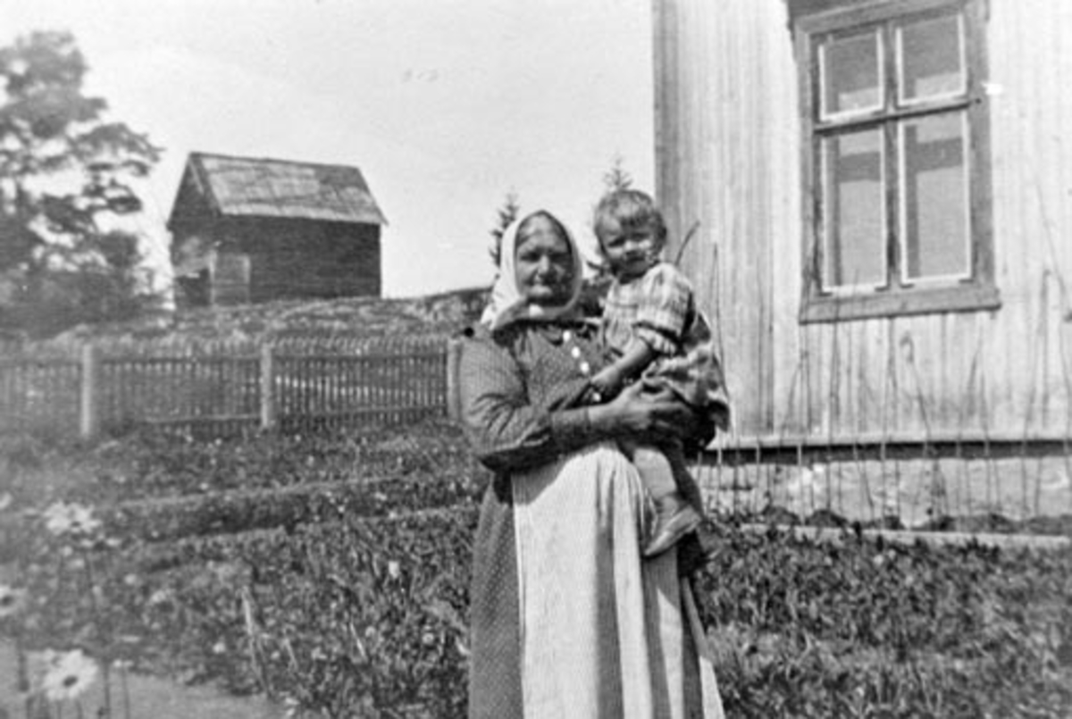 Anno Domkirkeodden, Dina Stenberg, vaskekone og bakstekone på Bjørnstad, Nes, Hedmark med Ingrid Feiring (1922-1946) på armen. I bakgrunnen det andre gamle stabburet som stod på gården. Dette er revet.