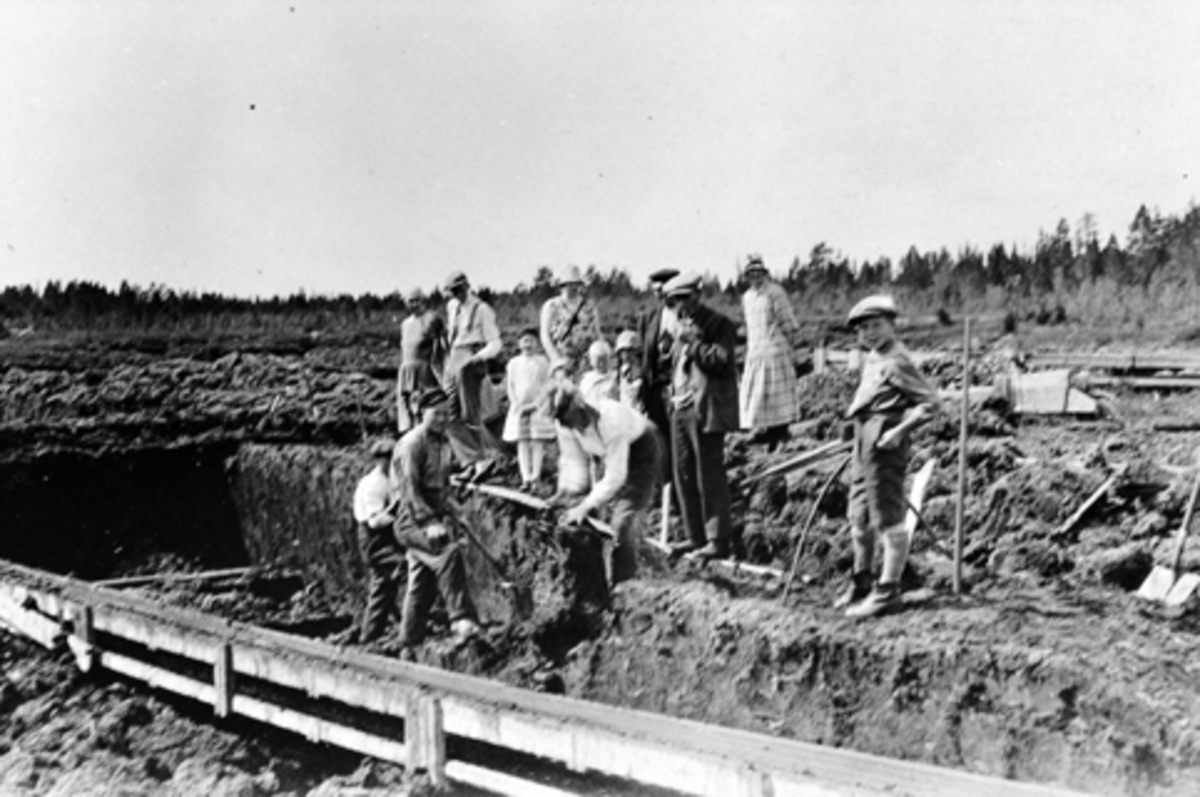 Anno Domkirkeodden, Voksne og barn arbeider på Leikmyra torvfabrikk. Brenntorvgruve i Furnes Almenning. Syverin Myhre, Egil Mikkelsen, Kåre Godager. Tilskuere er garver O. Kirkeby med familie, Petter Kirkeby med pipe og Erika Sveum. Gutten til høyre er Kristian Sveum.