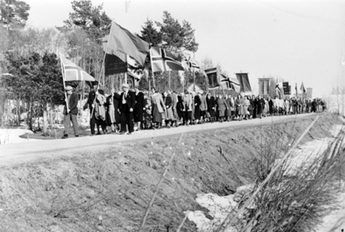 Anno Domkirkeodden, 1 mai-tog på Nes, Hedmark. Toget går fra Gaarum og skal til Folkets Hus på Stavsjø. Norske lagg og faner. Mannen som bærer den ensfargede fanen som nr. 2 i toget er Leon Teksum og til venstre for ham er Emil Elstad. Begge var sentrale personer i arbeiderpartiet og kommunepolitikken etter krigen.