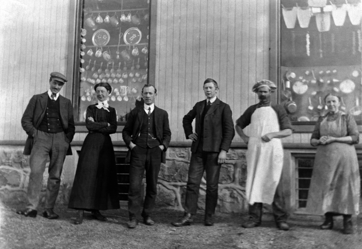 Anno Domkirkeodden, HØISTAD LANDHANDLERI, VINDUSUTSTILLING, GRUPPE: 6 FORAN FRA VENSTRE: LANDHANDLER OLE GJESTVANG, ASTRID GJESTVANG, HANDELSBETJENT KRISTIAN STENSBAK, OG JOHAN FUGLESANG, BAKER OPSAHL, HUSHOLDER MARIA, OTTESTAD.