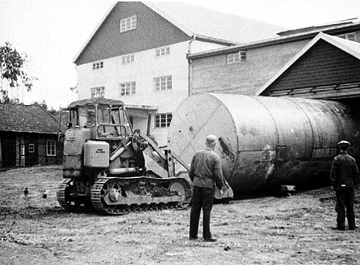 Anno Domkirkeodden, ATLUNGSTAD BRENNERI, BULLDOSER SKYVER SPRITTANK INN I NYTT LAGER, BULLDOSKJØRER: HELGE GJESTVANG, BRENNERIMESTER ERIK RØHNE OG RENSERIMESTER AKSEL OLSEN, PLASSERER SPRITTANKENE I DET GAMLE POTETLAGERET FRA 1955