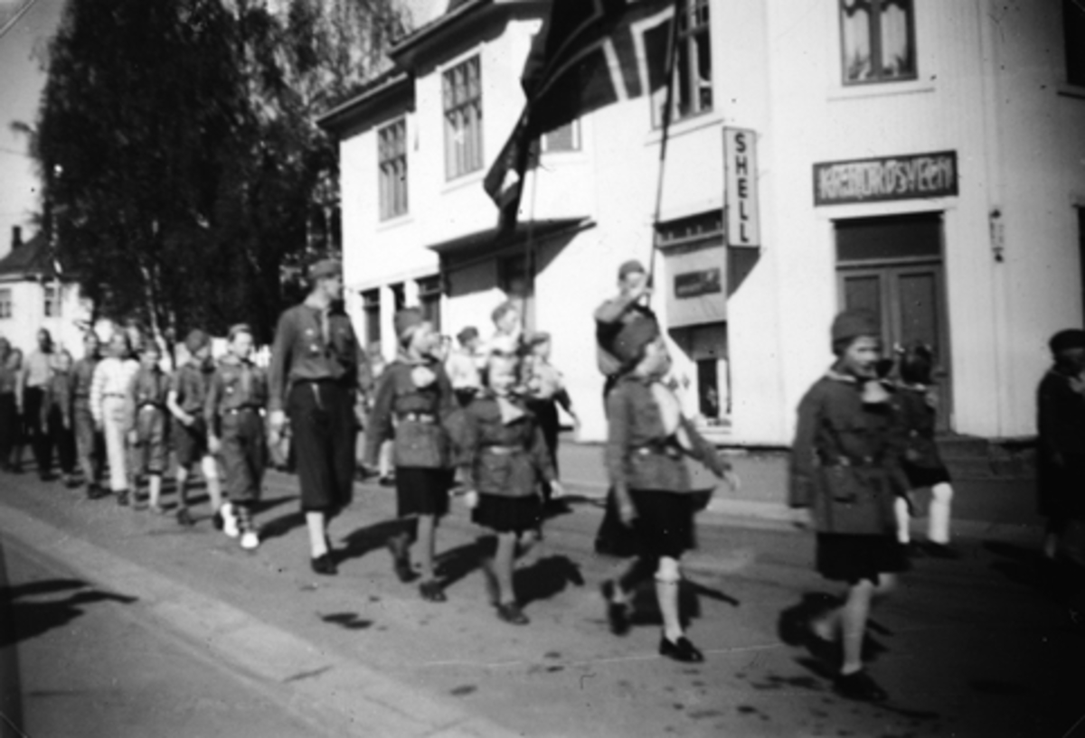 Anno Domkirkeodden, STORGATA I STANGE, SPEIDERTOGET PASSERER BUTIKKEN TIL NORDSVEEN 17. MAI CA. 1955. PERSONER SOM ER MED PÅ BILDET ER BLA. MAI TURID FINSTAD, ANNE KARI FARSTAD OG ANNE GRETHE JENSEN (MED HVITE STRØMPER. CA. 1955.