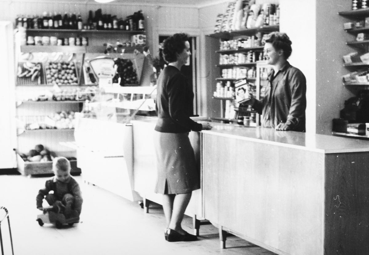 Ukjent, Rusten Landhandleri på Espa. 1960. Bak disken står Bodil Rusten f. Buenget. Foran Gunda Buenget og Ole Andreas Buenget sitter på gulvet og leker med lekebil. Butikkinteriør, kolonial Flere opplysninger se boka "Handelssteder i Stange" skrevet av Jan Arne Berg, utgitt av Stange Historielag 2012