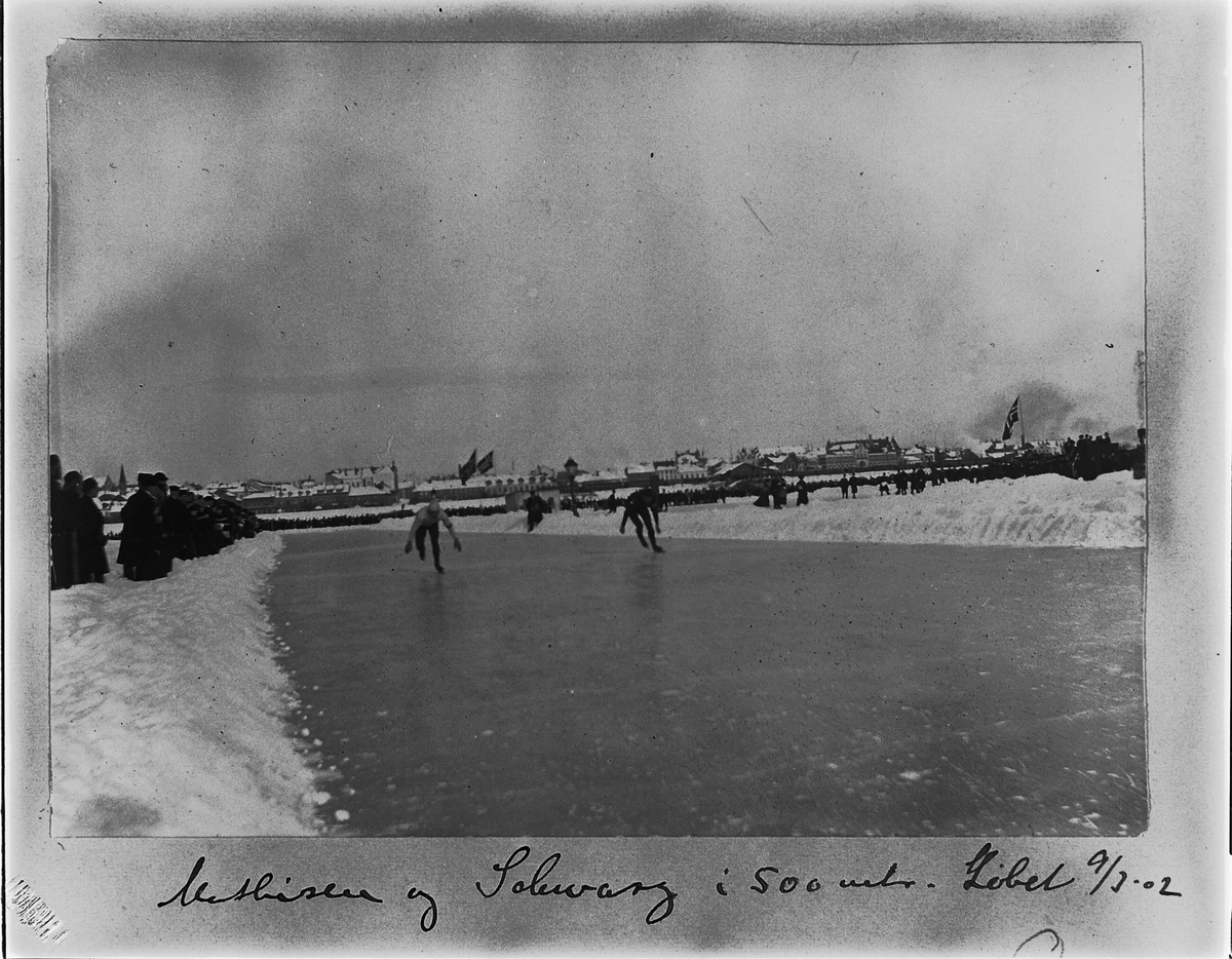 Johan Albert Steen, SKØYTELØP PÅ MJØSISEN, MATHISEN OG SOLVANG I 500 METERS LØPET, TRIBUNE, PUBLIKUM, FLAGG, 9-3-1902, BEBYGGELSEN I STRANDGATA, HAMAR I BAKGRUNNEN.
