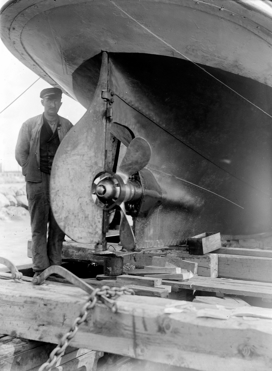 Oscar Løberg, MJØSBÅT, MANN VED PROPELL OG ROR, PÅ BEDDINGA RETT UTAFOR EKELY PÅ STERUDODDEN VED HAMAR, MANNEN KAN VÆRE VOGNMANN OLA OLSEN. 1922. Dette er "Bundefjord". Sommeren 1922 ble dampmaskina byttet ut med motor. Samtidig byttet de propellen. Den nye propellen var så stor at det måtte skjæres ut en bit av…