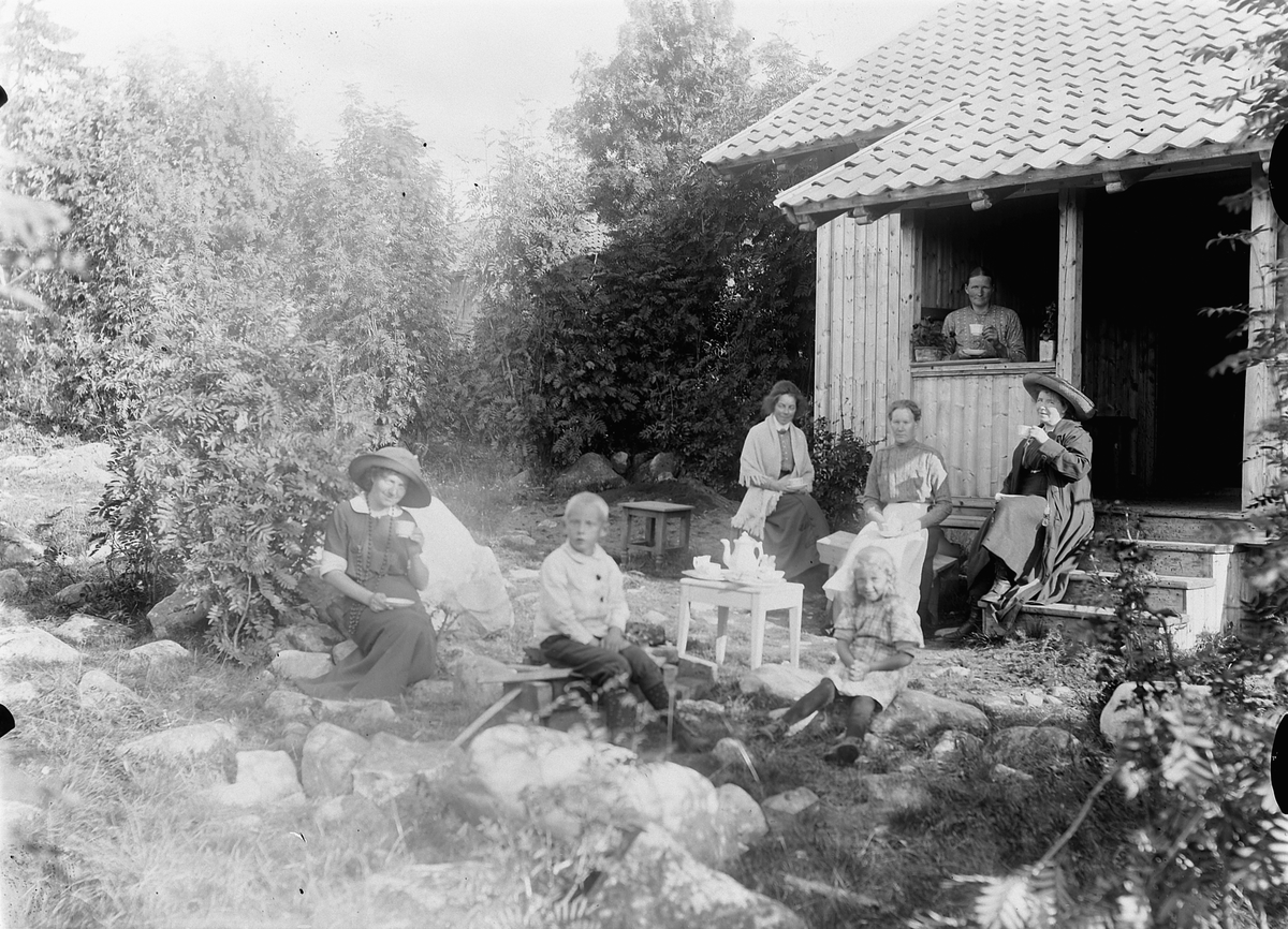 Mina Skraastad, Gruppe kvinner, og to barn, drikker kaffe utenfor et hus med hage.