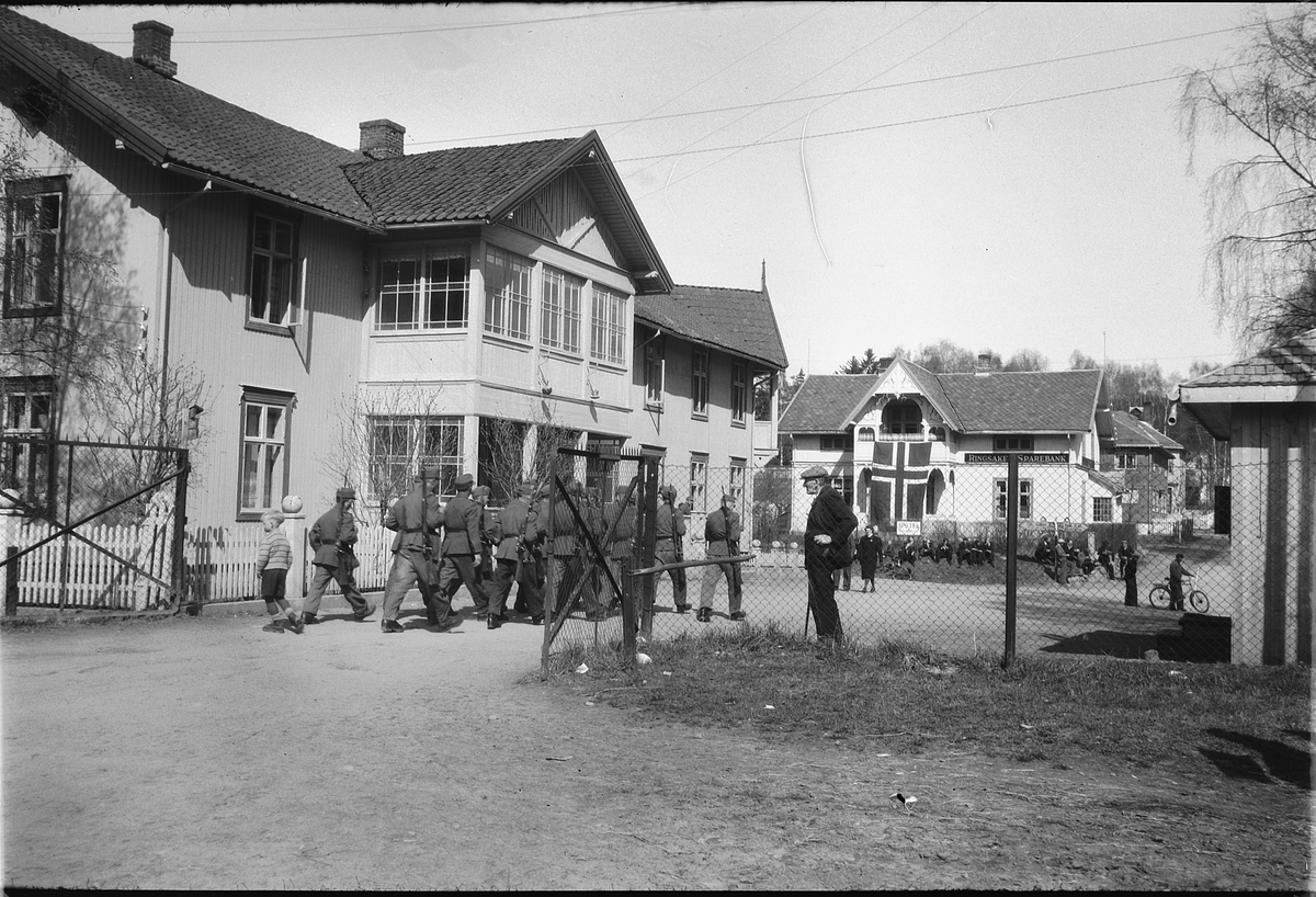 Håkon Prestkværn, Gruppe soldater med uniform og våpen marsjerer inn en port ved Ringsaker Sparebank, Storgata i Moelv, fredsdagen etter 2. verdenskrig.