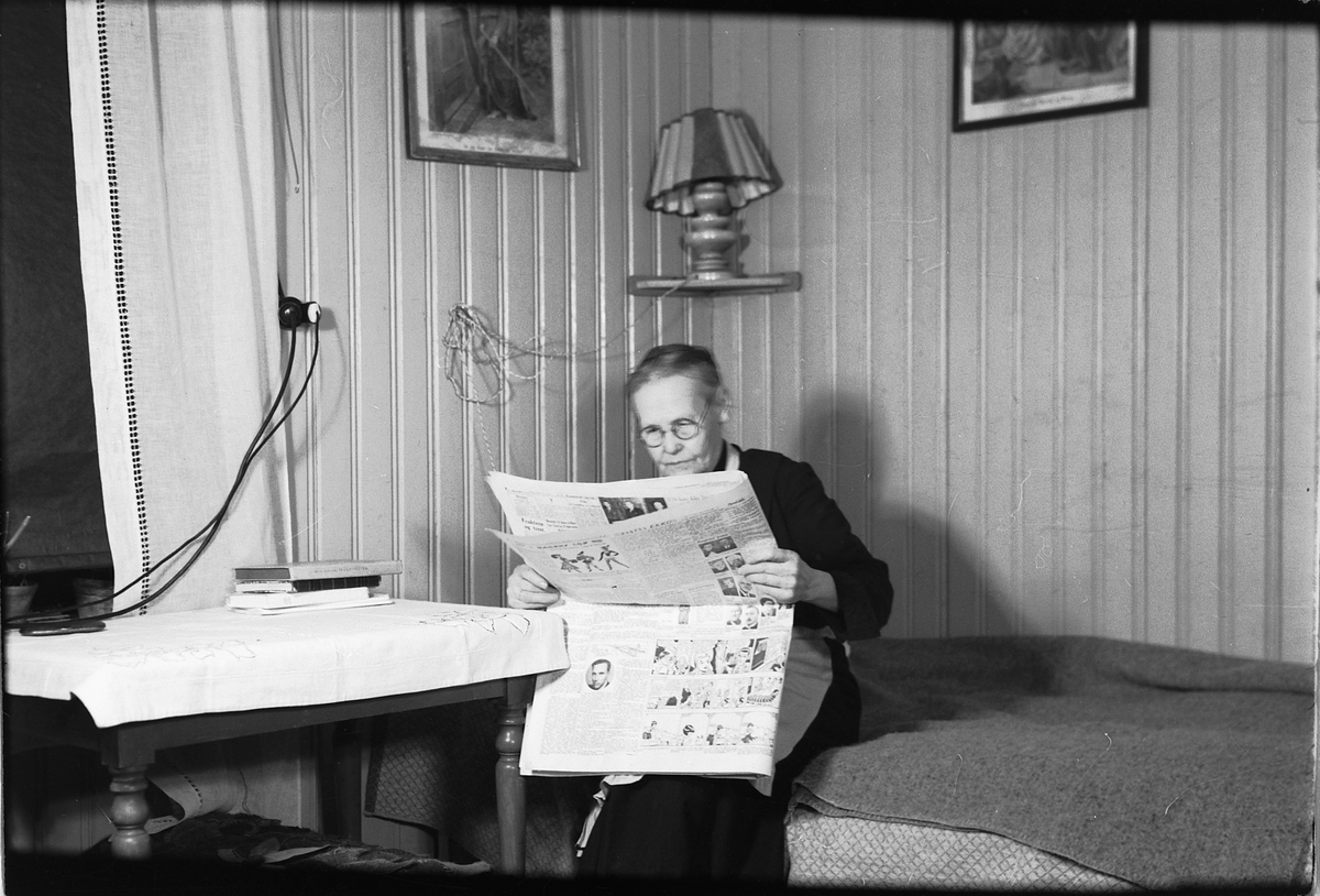 Håkon Prestkværn, Interiør, stue. Eldre kvinne sitter på sofaen og leser ei avis.