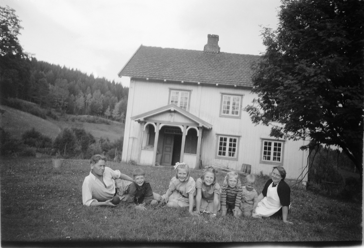 Håkon Prestkværn, Ukjent kvinne og mann med en gruppe barn i gresset foran et hus.