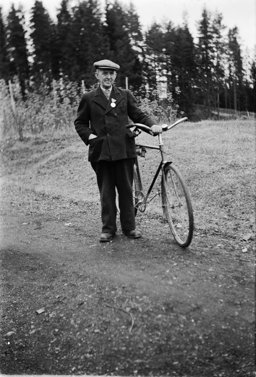 Håkon Prestkværn, Eldre mann med sixpence og medalje på jakkekragen holder en sykkel på veien.