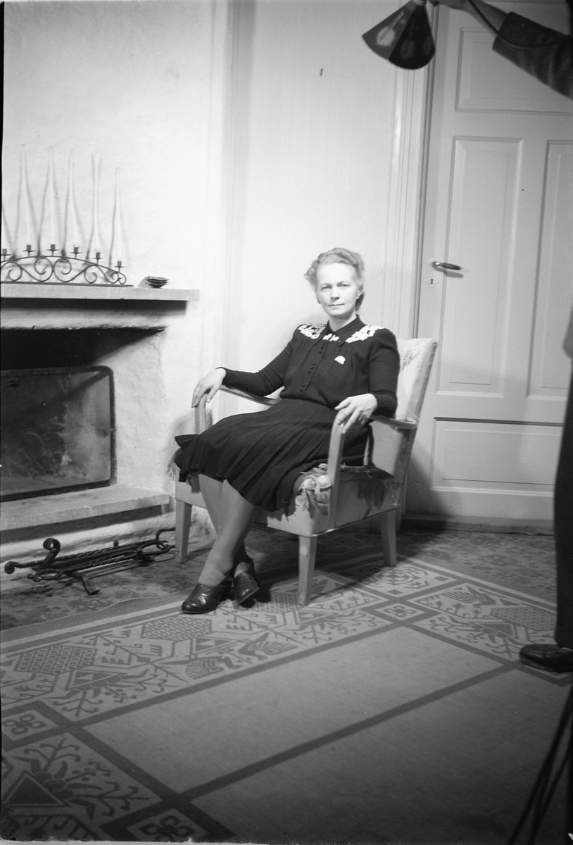 Håkon Prestkværn, Interiør, stue. Kvinne sitter i en stol ved peisen.
