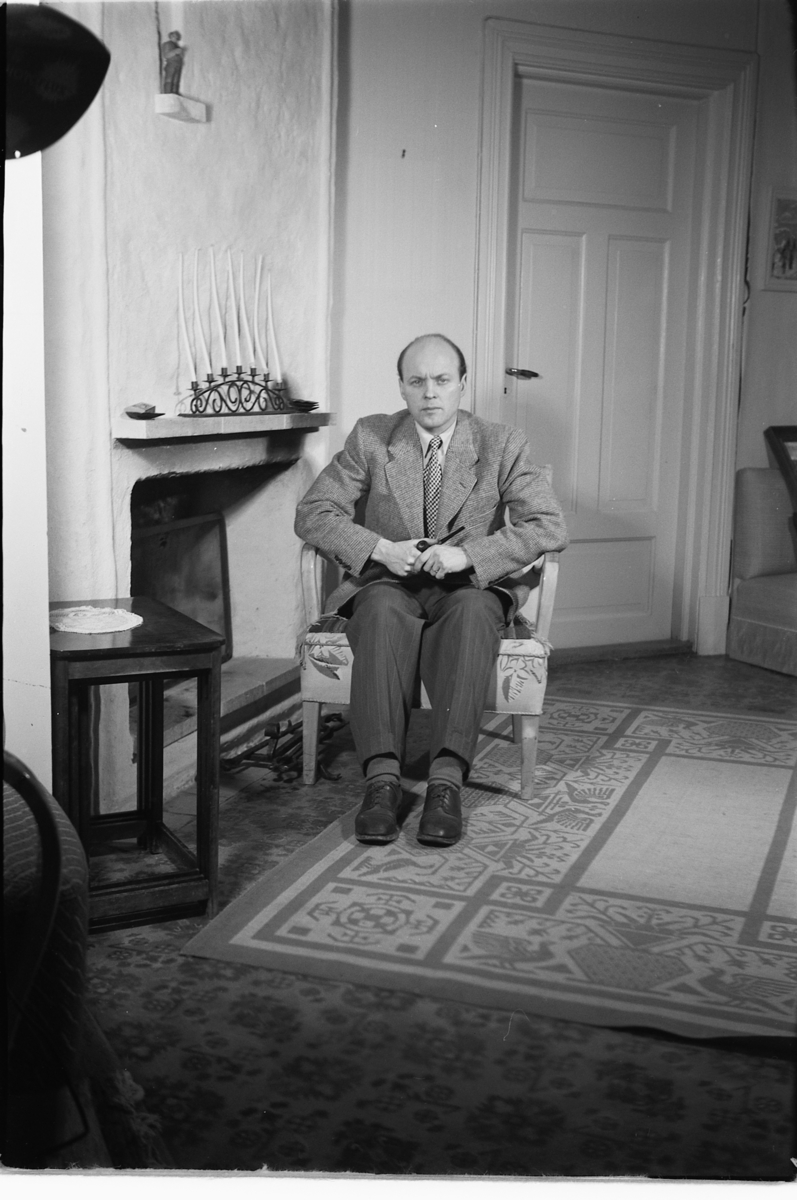 Håkon Prestkværn, Interiør, stue. Ukjent mann sitter i en stol ved en peis.