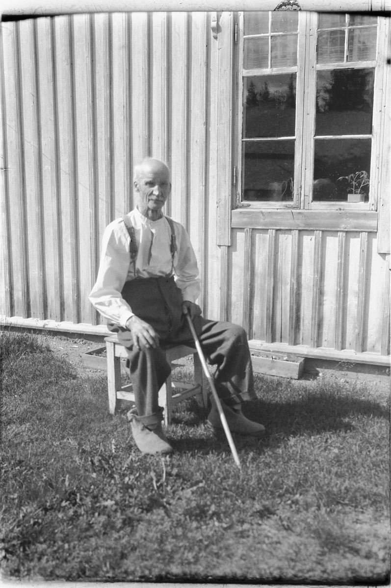 Håkon Prestkværn, Eldre mann med stokk sitter på en krakk ved husveggen.