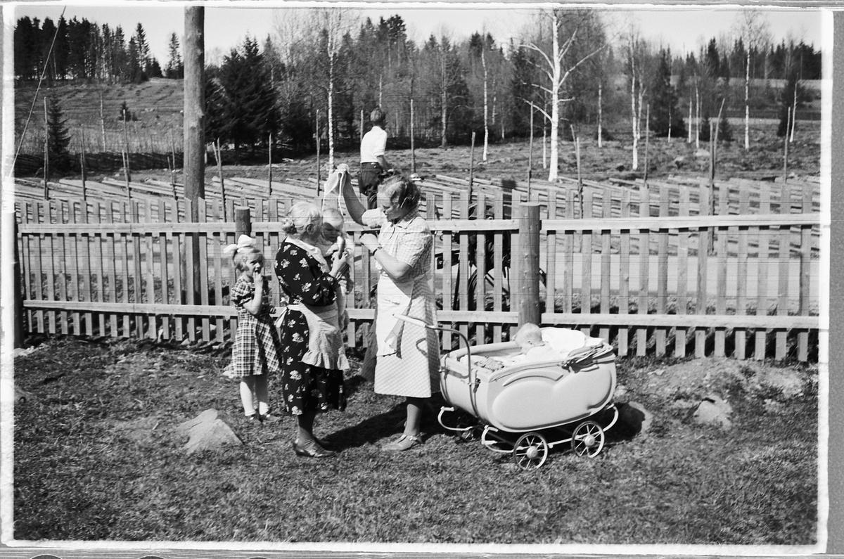 Håkon Prestkværn, Ukjent familiegruppe med to kvinner med en baby på armen, ei jente, og en baby i barnevogn. Tvillingene er Knut og Bjørg Svenskerud f. 1949. Jenta med sløyfe er Inger Svenskerud f. 1945. Det ser ut til å være mor Aslaug født Kindsbekken som hjelper med påkledning. Bildet er tatt ved Åsmarka kirkest…
