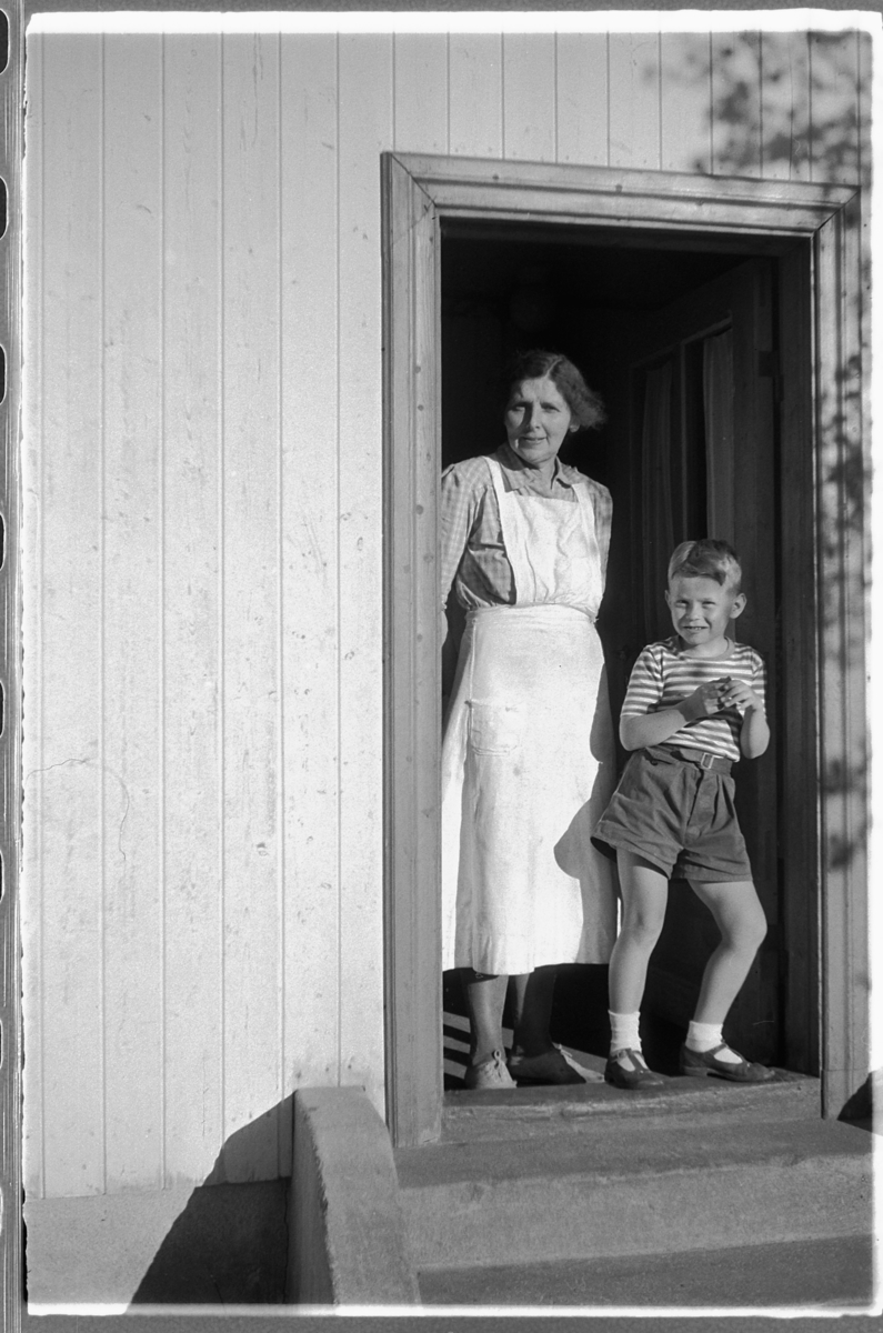 Håkon Prestkværn, Ukjent kvinne med forkle sammen med gutt i inngangsdøra til et hus.