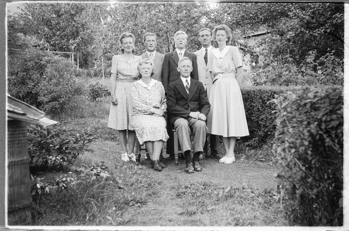 Håkon Prestkværn, Eksteriør, hage, familiegruppe. Foran sitter Palma, født Bjerke fra Brøttum, og Robert Mostue (1896-1997). Sammen bygget de hjemmet Bjerke i Jenstuvegen, Moelv. Bak står Anna Lilli f. 1919 og ektemann Einar Lødding fra Andenes f. 1916, Leif Mostue f. 1934, Leif Hagen fra Oslo og kone Arna Kristine …