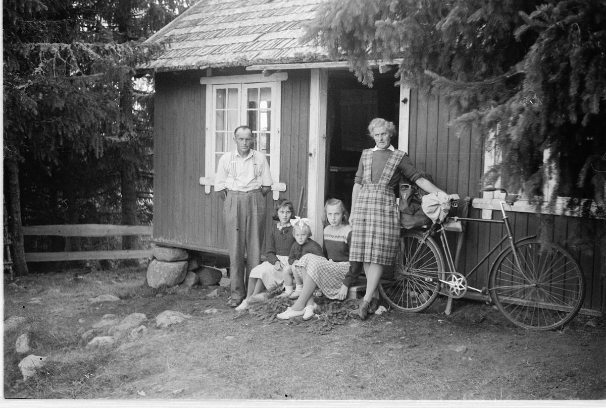 Håkon Prestkværn, Ukjent mann, kvinne og tre jenter ved inngangsdøra til et hytte. Kvinnen holder i en sykkel. Ljøsheim?