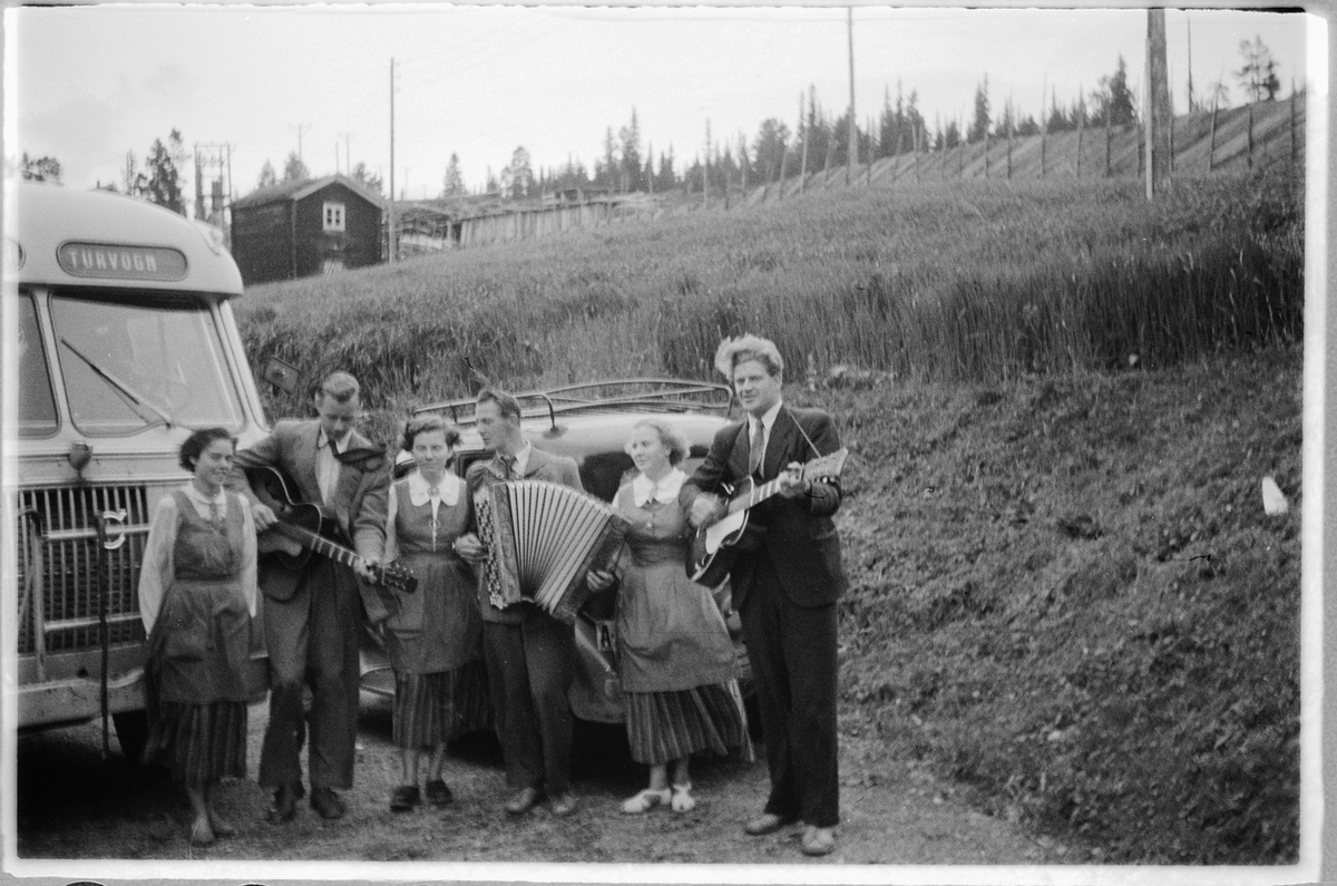 Håkon Prestkværn, Tre menn spiller gitar og trekkspill, mens tre kvinner lytter ved en buss med skilt Lillehammer turvogn. T. h. Ulf Sæhli på gitar. Se HHB-7409-28.