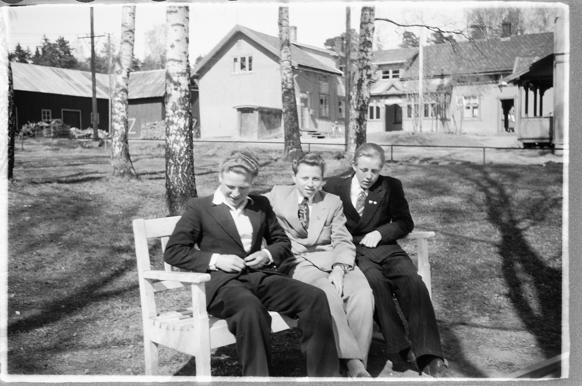 Håkon Prestkværn, Tre unge menn, i dress sitter på en benk. I midten Reidar Larsen (Baashus), de to andre er ukjente. Kan alle tre være lærlilnger for Frisør Johansen? I bakgrunnen Fruseth kafeen.