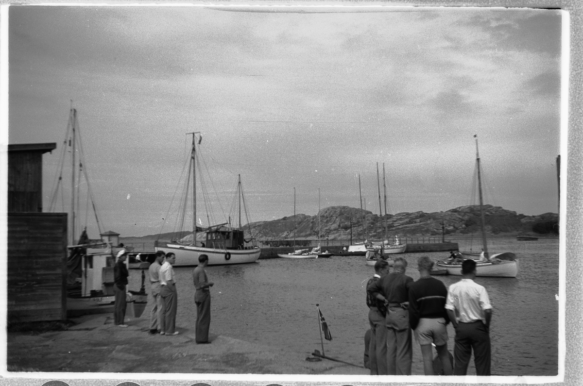 Håkon Prestkværn, Gruppe mennesker og båter ved en havn i en skjærgård.