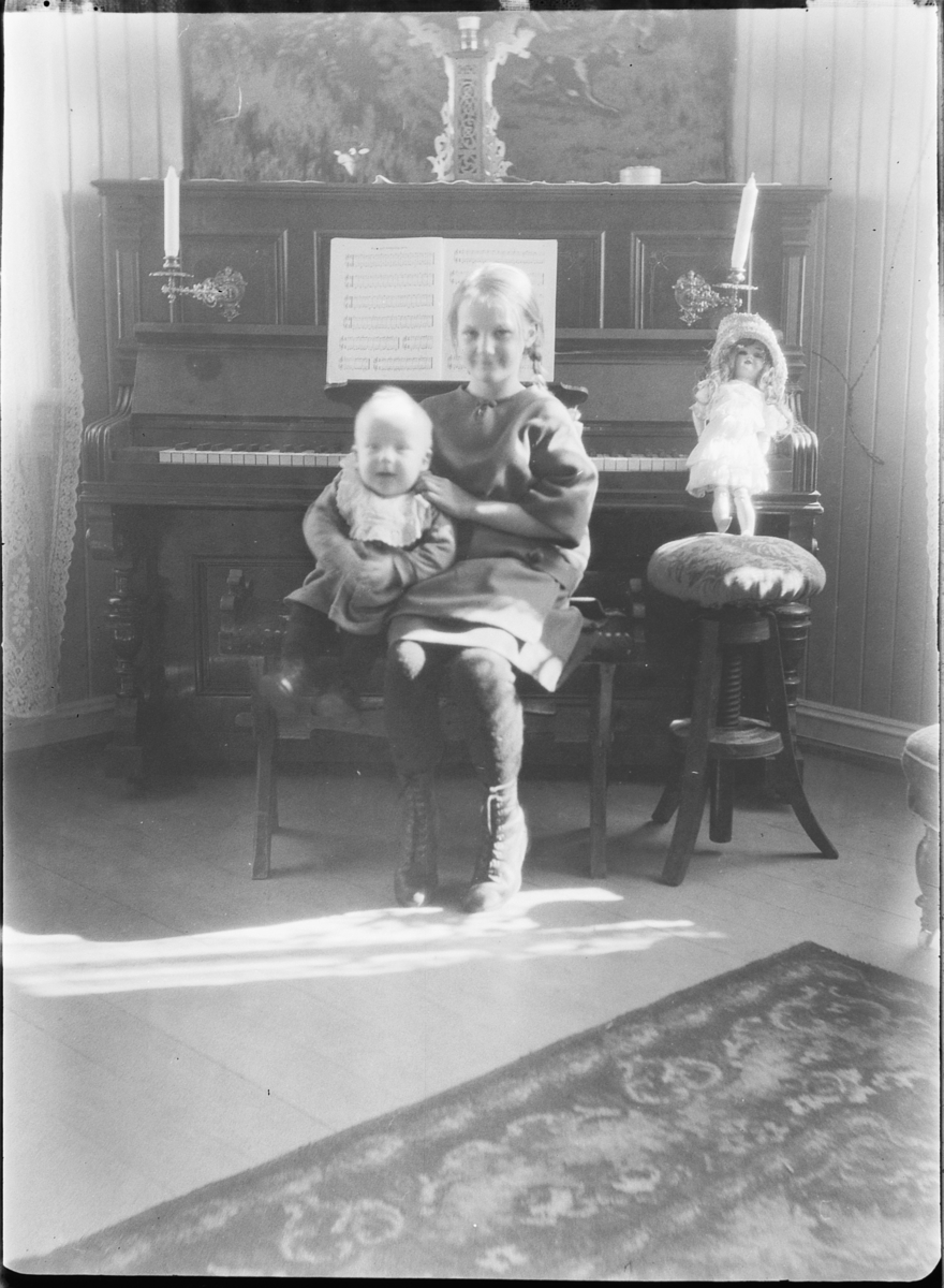 Ole M Martinsen, Interiør, stue. Jente med en baby på fanget sitter foran et piano og en dukke.