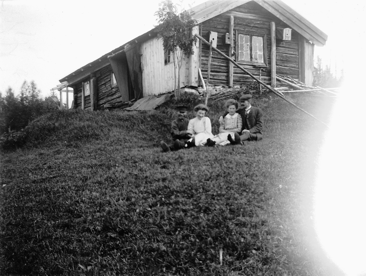 Ragnvald Andersen, To par sitter på gresset foran tømmerbygning. Seterhus? Ukjent. (Ragnvald Andersen til høyre?)