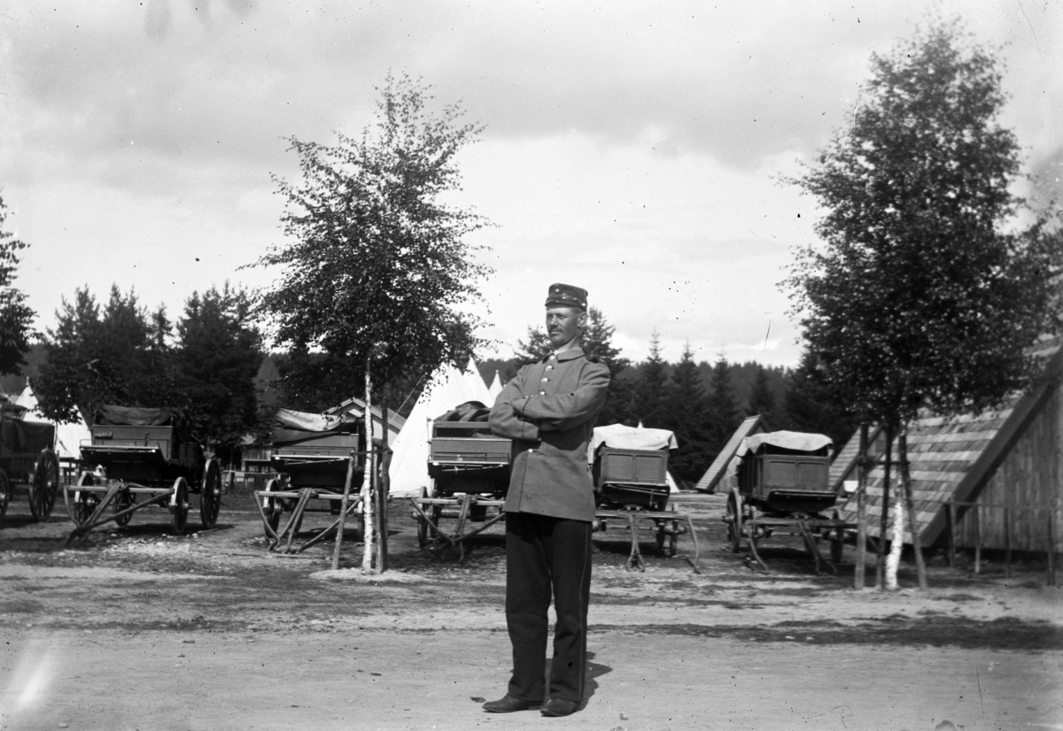 Johannes Larsen Alhaug, En soldat i uniform foran ei gruppe kjerrer, telt i bakgrunnen.