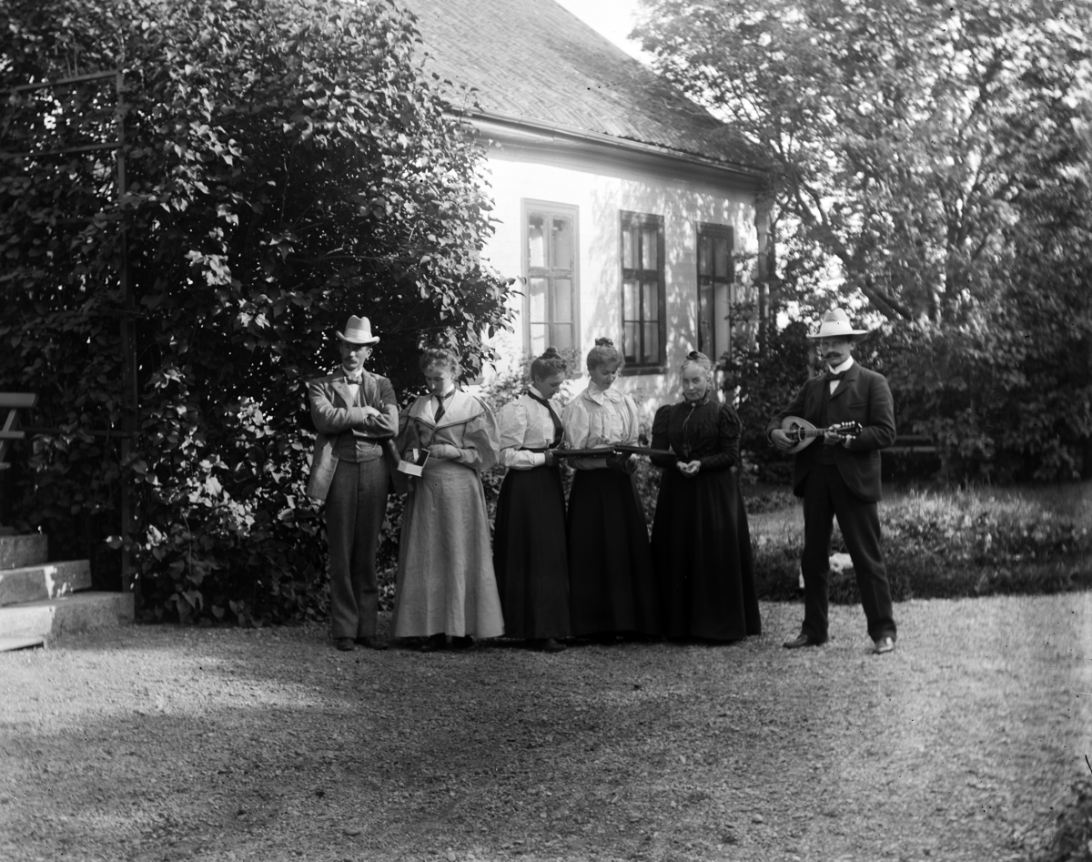 Martin Finborud, Gruppe kvinner og menn i en hage. Mannen til høyre spiller på en mandolin. Mellbye, Grefsheim, Nes, Hedmark.