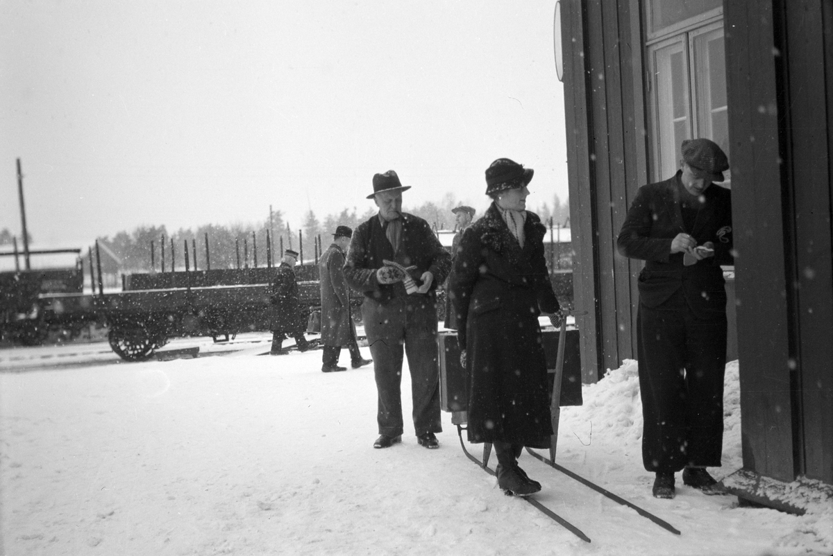 Håkon Prestkværn, Moelv jernbanestasjon, Vinter, snøvær. Dame med spark og koffert, ukjente.