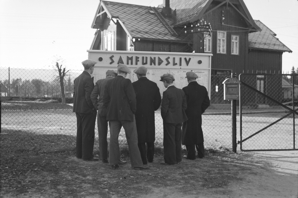 Håkon Prestkværn, Moelv jernbanestasjon. Ungdom studerer oppslagstavle "Samfundsliv" Postkasse henger på gjerdet. T. h på gjerdet henger postkasse.