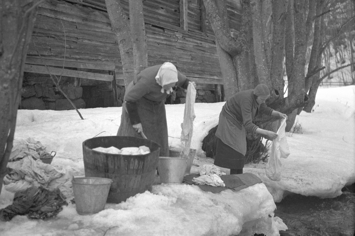 Håkon Prestkværn, Klesvask ved den gamle mølla til Prestkværn i Moelva, vinter, klesstamp, sinkbøtter. Vaskekoner ved Moelva. T. v. Mina Lunna. Ca. 1938. På iskanten om vinteren skylles og vris tøyet. Før vannverket kom var dette et vanlig syn. Langs elvebredde sto også gryter og tønner som klærne ble kokt i og ette…