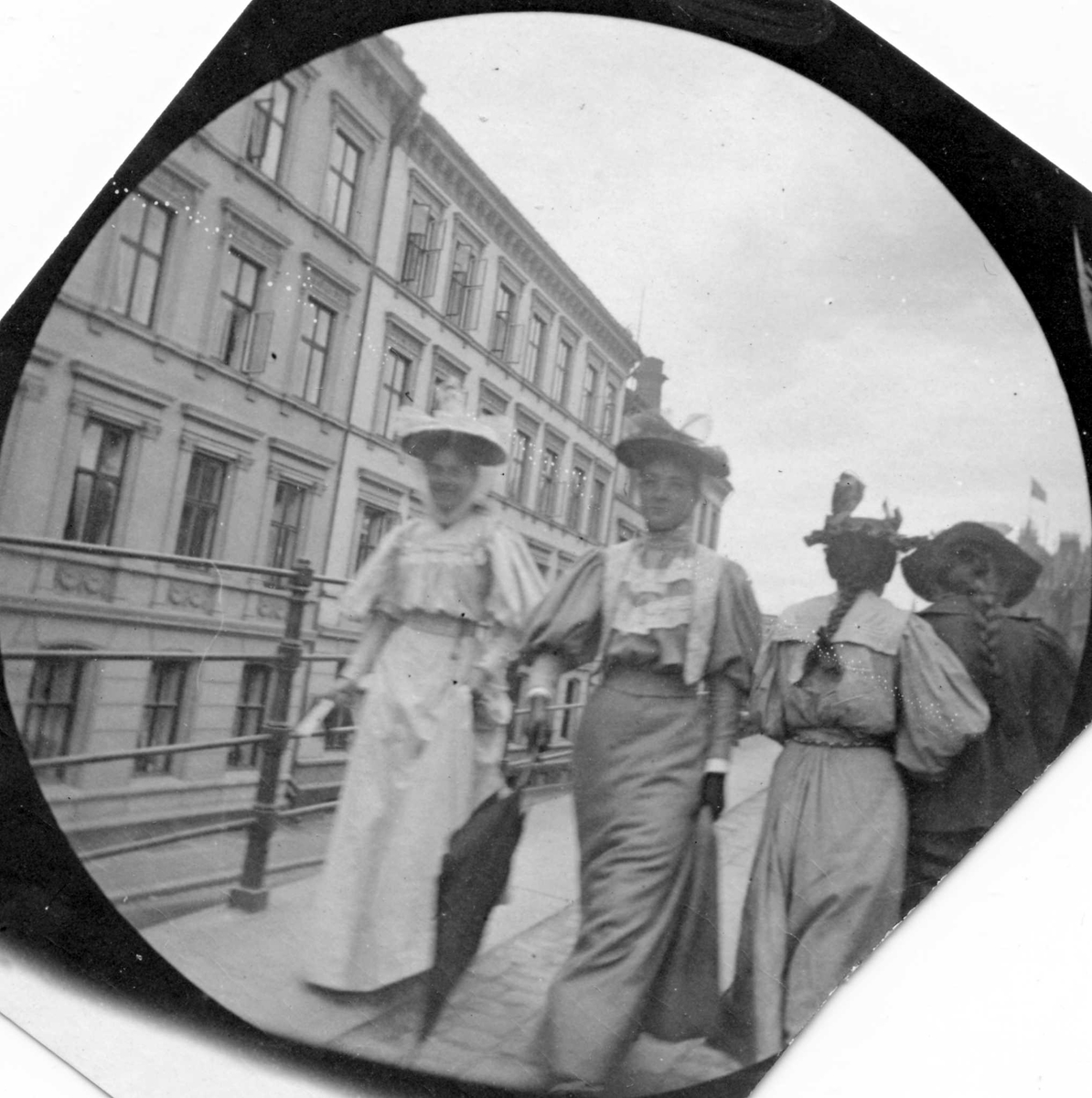 Fredrik Carl Mülertz Størmer, Frk.Bjelke og frk. Hvoslef spaserer langs gjerde i bygate, Oslo. To damer med ryggen til og bygårder.