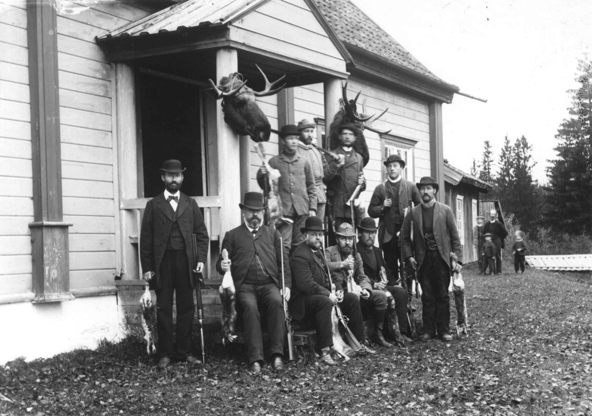 Fredrik Emil Faye, Gruppeportrett, jaktselskap for harejakt, mannsgruppe med våpen og bytte utenfor hus. Elghoder hengt opp ved inngangen. Fra serie fotografert av kammerherre Fredrik Emil Faye (1844-1903), Dal gård, Ullensaker.