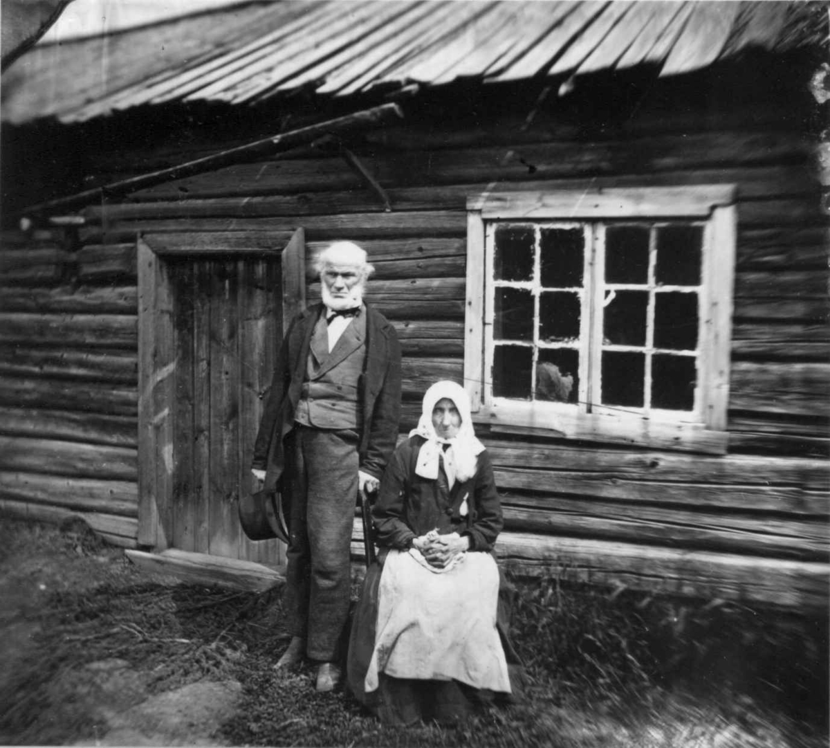 Fredrik Emil Faye, Husmannsfolk, Dal gård, Ullensaker. Eldre par poserer utenfor huset sitt, ca. 1879. Fra portrettserie av personer som bodde på eller besøkte Dal gård, Ullensaker, fotografert av gårdens eier, kammerherre Fredrik Emil Faye (1844-1903) i årene 1875-1900.