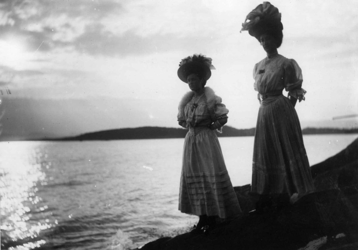 Hermann Christian Neupert, Drakt. Oslo 1908 - 1910? Fjordlandskap? I forgrunnen to kvinner i lange kjoler. Bærer hatter.